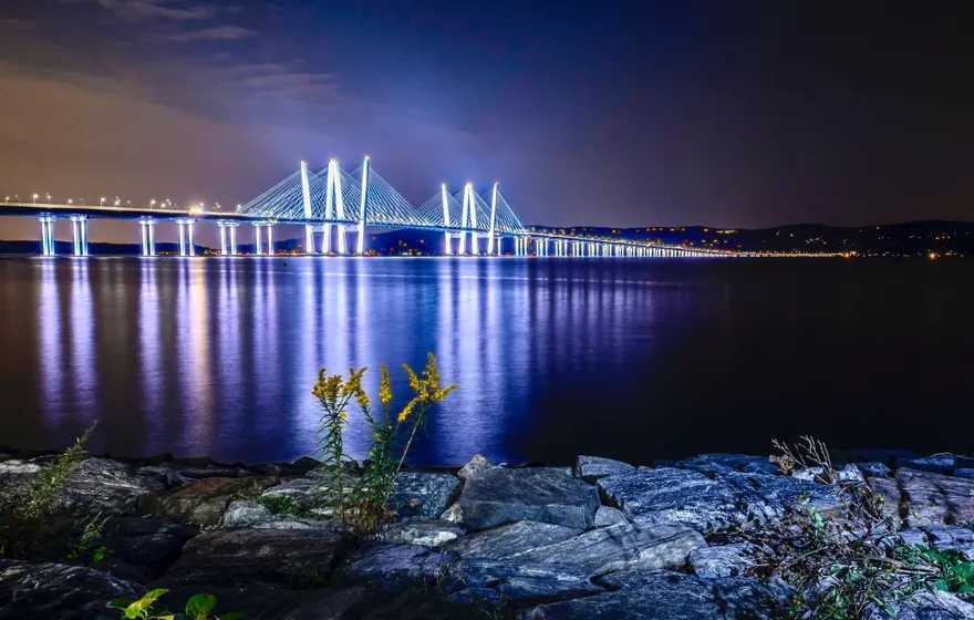 A long cable-stayed bridge lit blue and white at night over the Hudson River, its lights reflected in the water, rocks and wildflowers on the shore in front