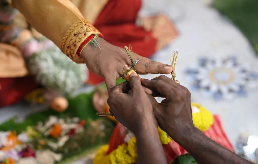 Close-up of one person's hands tying a yellow thread with a sprig of grass around another person's wrist, flowers and leaf plates of offerings on the floor below