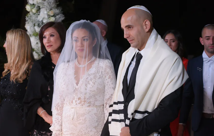 A bride in a lace gown and a sheer veil over her face stands beside a groom in a white kippah with a striped tallit over his black suit, guests standing behind them