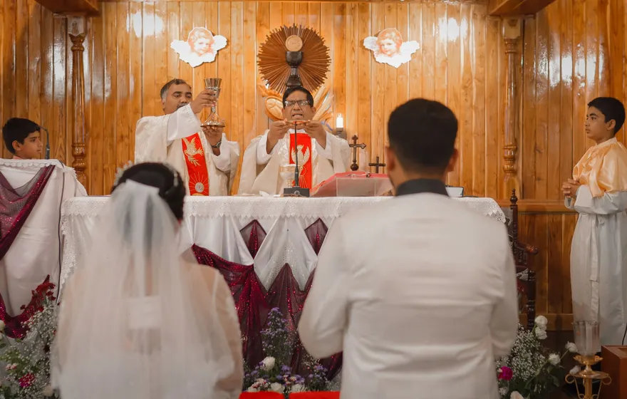 Priests in red and white vestments at a wooden church altar, one raising a chalice, while a veiled bride and a groom in a white jacket kneel in front of them