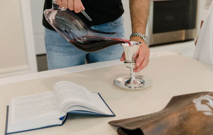 Red wine poured from a glass decanter into a small silver cup on a table beside an open book