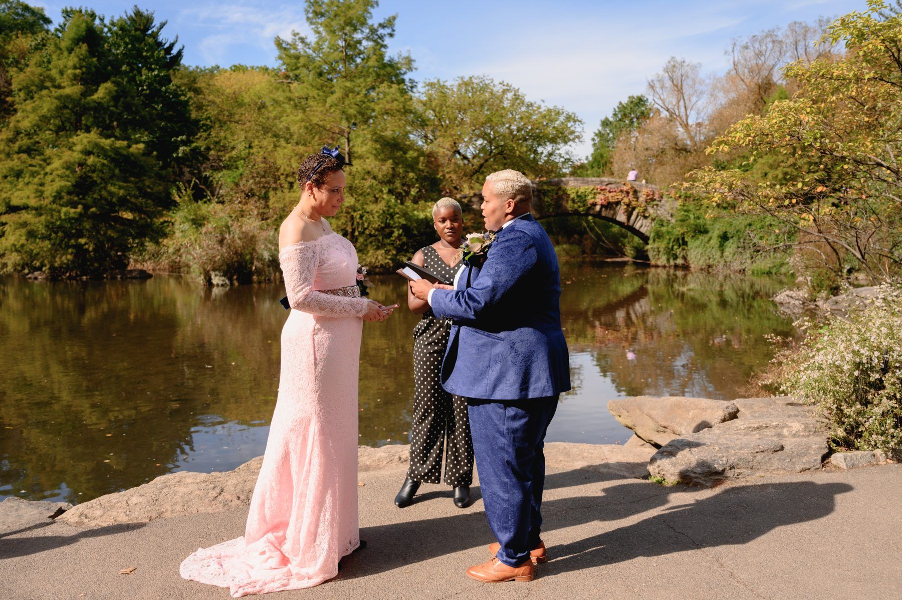 Two partners exchanging vows beside the water at Gapstow Bridge in Central Park while Robyn holds the script