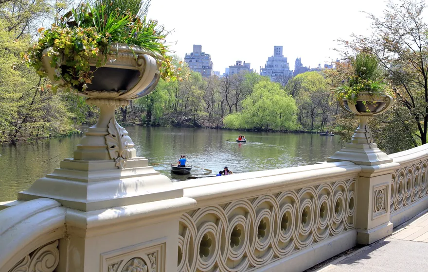 The cream cast-iron railing of Bow Bridge with its circular openwork and a planted urn on the post, rowboats on the Lake and the skyline beyond