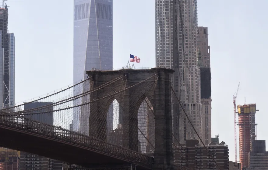 The stone tower and cables of the Brooklyn Bridge seen from Empire Fulton Ferry Park, One World Trade Center rising behind it
