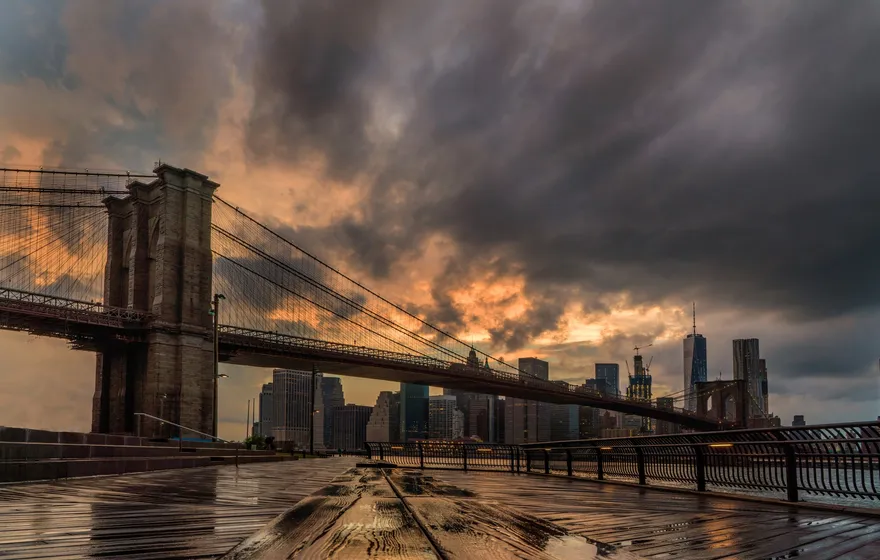 The Brooklyn Bridge and the Lower Manhattan skyline at sunset under dramatic orange and gray clouds, seen from a wet boardwalk in Brooklyn Bridge Park