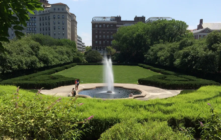 The jet fountain in the Center Garden of the Conservatory Garden, framed by clipped hedges, with apartment buildings on Fifth Avenue behind the trees