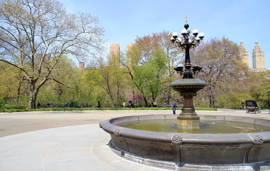 The Cherry Hill fountain in Central Park on its paved plaza in spring, trees just leafing out and the twin towers of the San Remo beyond