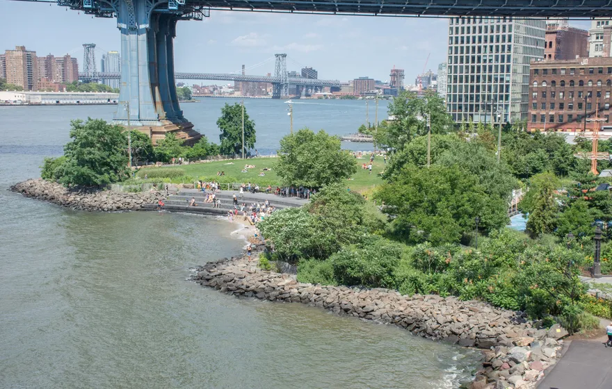 An aerial view of the Main Street section of Brooklyn Bridge Park, a curved rocky shoreline and small beach on the East River beneath the Manhattan Bridge
