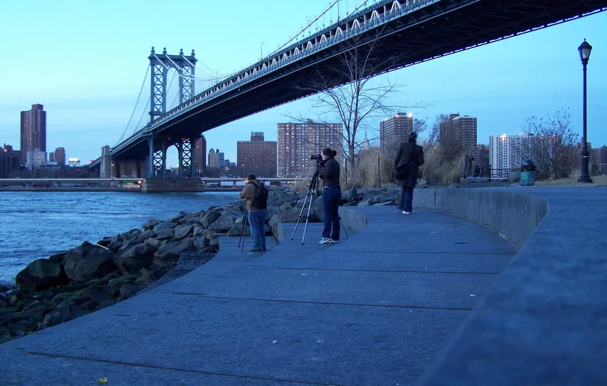 Photographers with tripods on the stone waterfront path beneath the Manhattan Bridge in Brooklyn at dusk, the bridge's steel span overhead