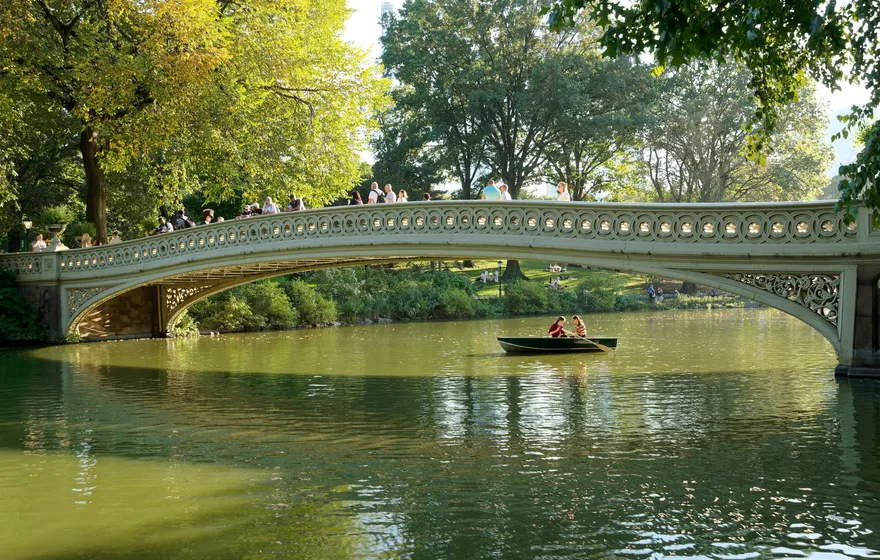 Bow Bridge spanning the Lake in Central Park on a summer afternoon, people standing and walking along its railing while a rowboat passes underneath