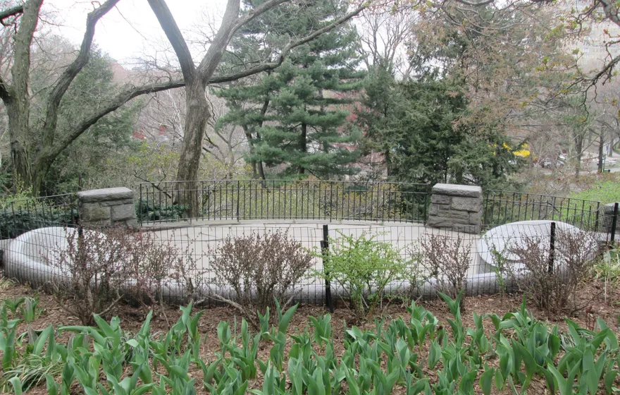 A low curved stone bench behind an iron railing in Shakespeare Garden in early spring, tulip leaves coming up in the bed in front
